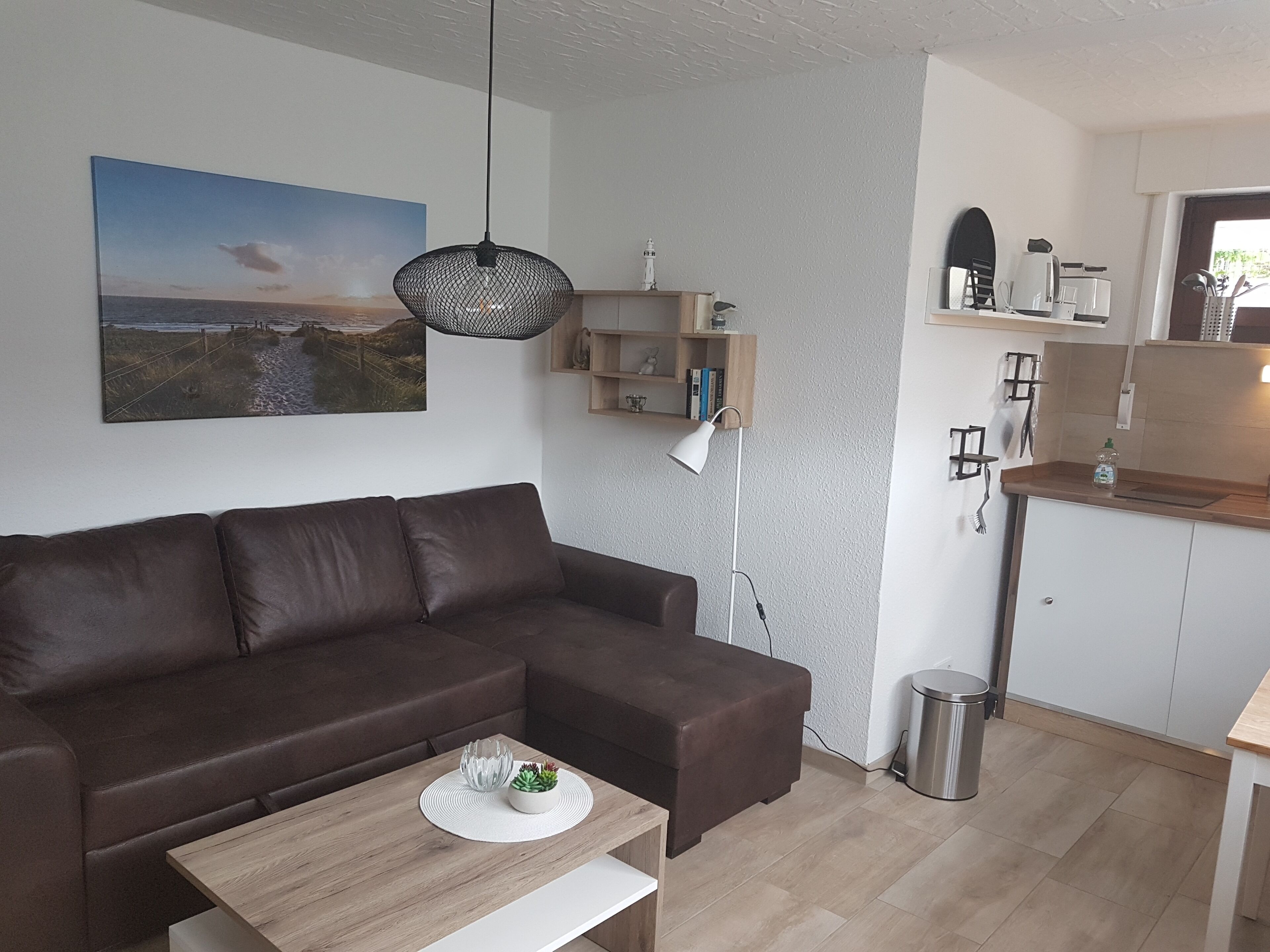 Een moderne zithoek met een bruine leren bank, een houten salontafel en een witte bijzettafel. Op de muur hangt een schilderij van een strandtafereel, er is een hanglamp en een klein plankje met boeken en decoratieve objecten.