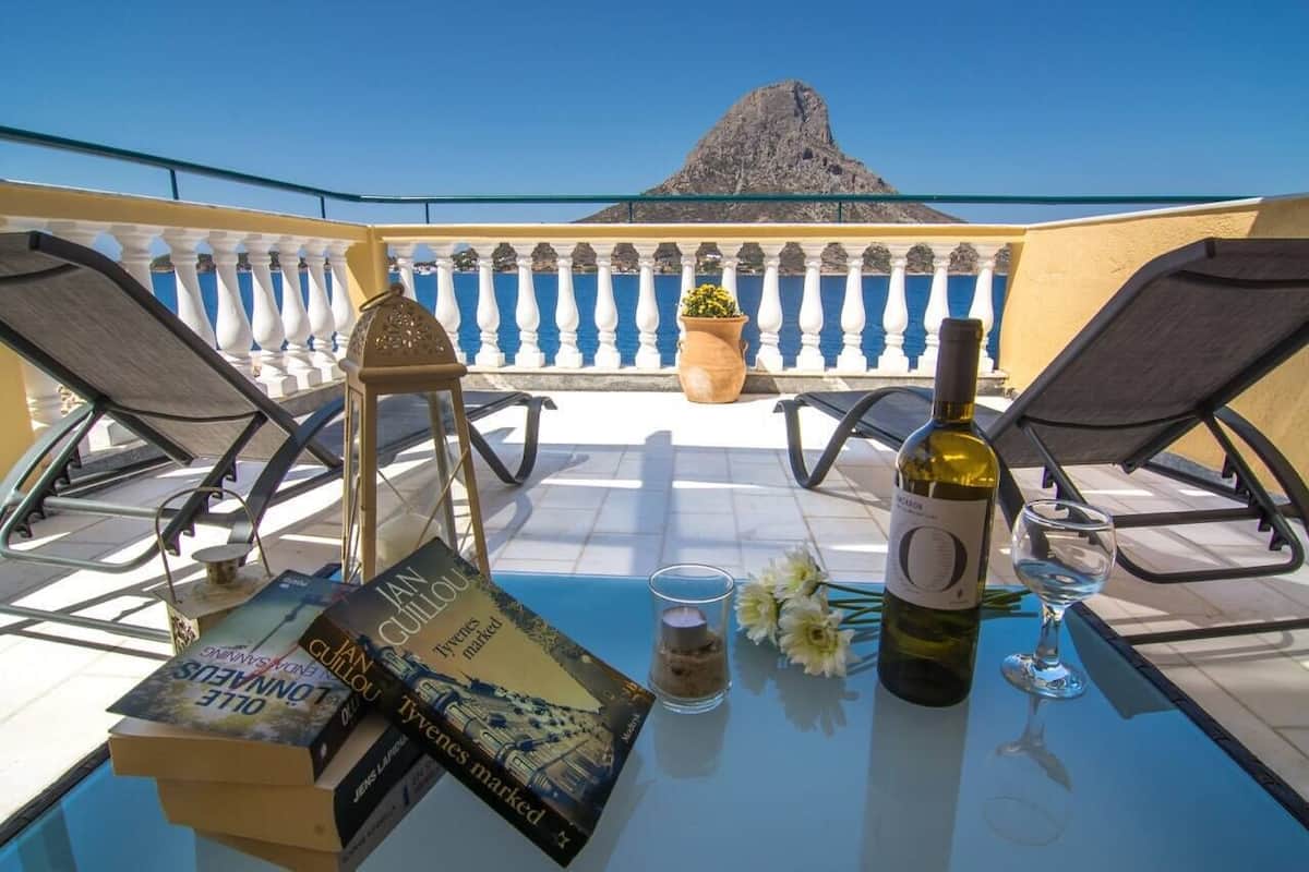 A table with a book, a wine bottle, and a glass on a balcony overlooking a mountain and sea.