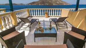 A terrace with wicker furniture, a glass table, and a view of a mountain and sea. - Elena Apartments (Kalymnos)