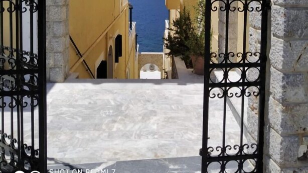 An entrance gate with the name "ELENA" carved into a stone archway, leading to a pathway flanked by yellow buildings and a mountain in the background. - Elena Apartments (Kalymnos)