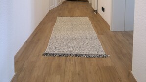 Ein Flur mit Holzboden, weißen Wänden und einem großen Teppich. Am Ende befinden sich zwei Holztüren, ein Heizkörper und ein gerahmtes Bild an der Wand.