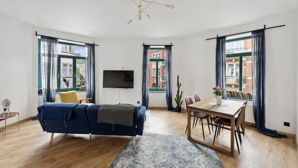 Ein modernes Wohnzimmer mit einem blauen Sofa, einem Esstisch mit Stühlen, einem Flachbildfernseher und großen Fenstern mit Vorhängen.