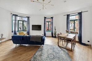 Un salon moderne avec un canapé bleu, une table à manger avec des chaises, un téléviseur à écran plat et de grandes fenêtres avec des rideaux.
