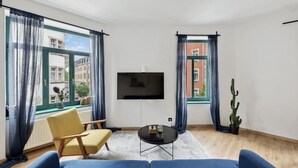 Ein modernes Wohnzimmer mit einer Couch, einem gelben Sessel, einem Couchtisch und einem an der Wand montierten Fernseher. Zwei Fenster mit Vorhängen, eine Kakteenpflanze und ein Blick auf Gebäude im Freien sind zu sehen.