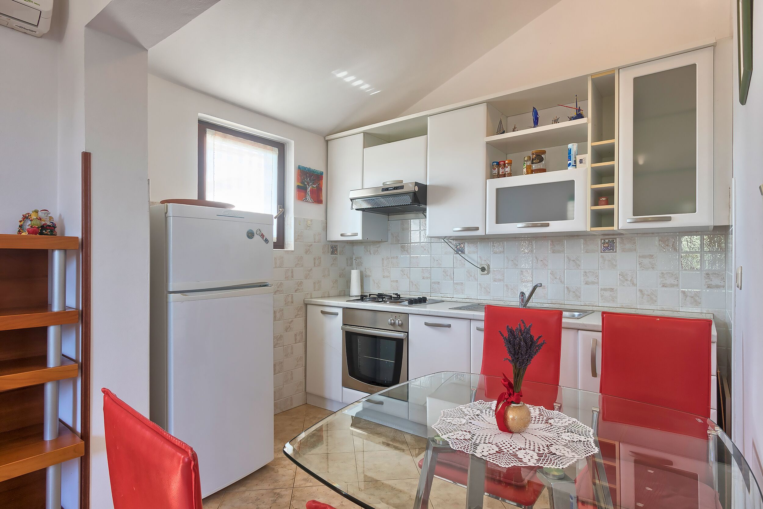 Una cucina moderna con mobili bianchi, un frigorifero in acciaio inossidabile, un fornello a gas e un tavolo da pranzo in vetro con sedie rosse.