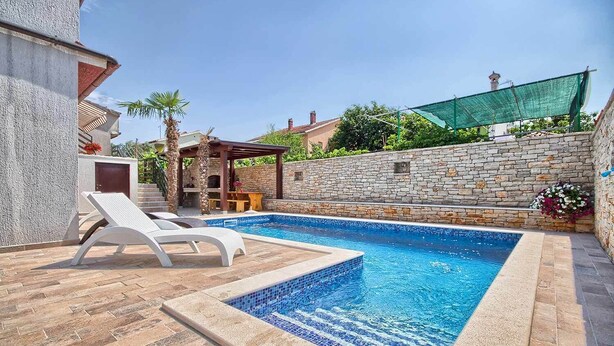Una piscina rectangular con borde de azulejos, rodeada por un muro de piedra y una pérgola.