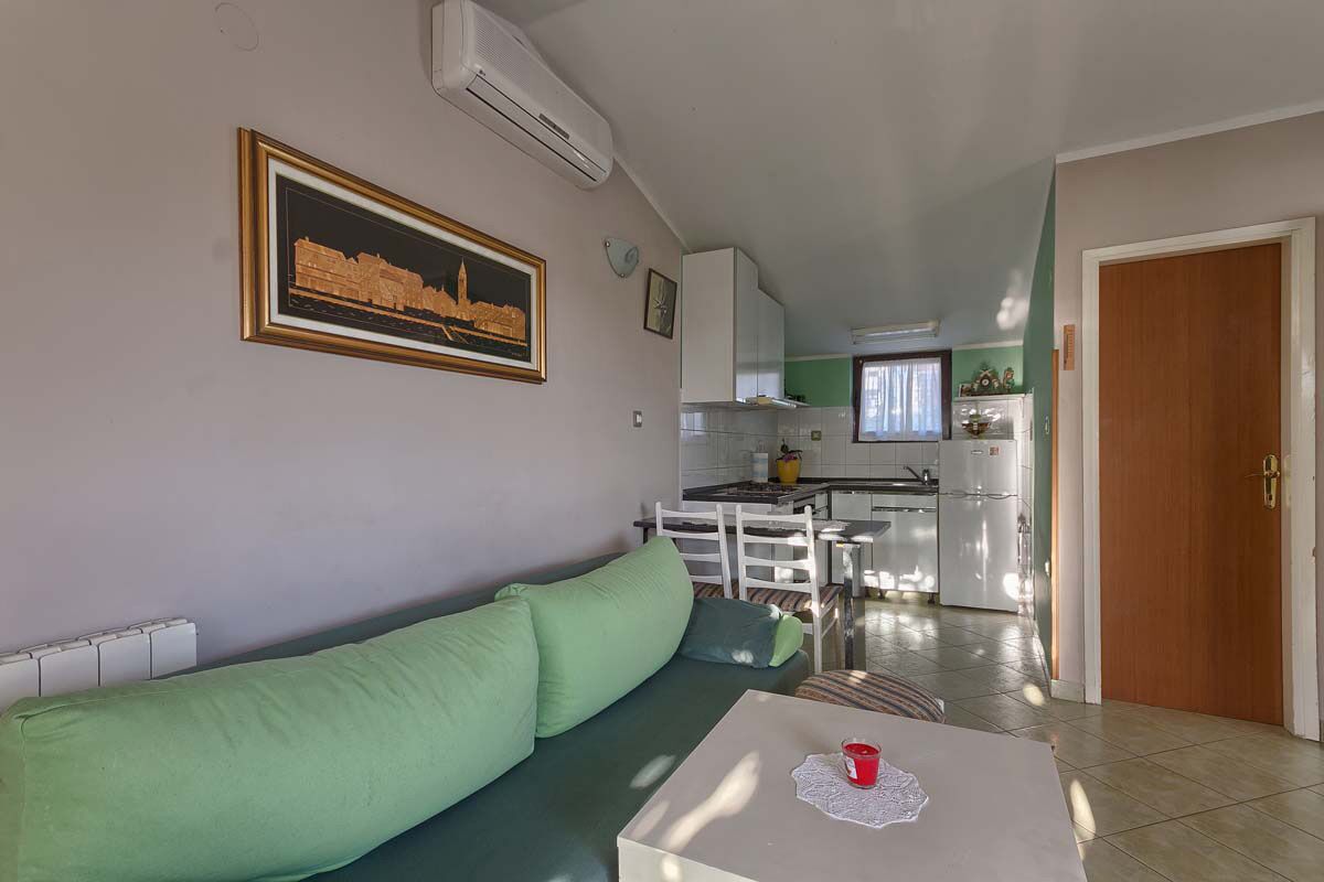Una sala de estar con un sofá verde, una pequeña mesa con una taza roja, un cuadro enmarcado en la pared y una cocina al fondo.