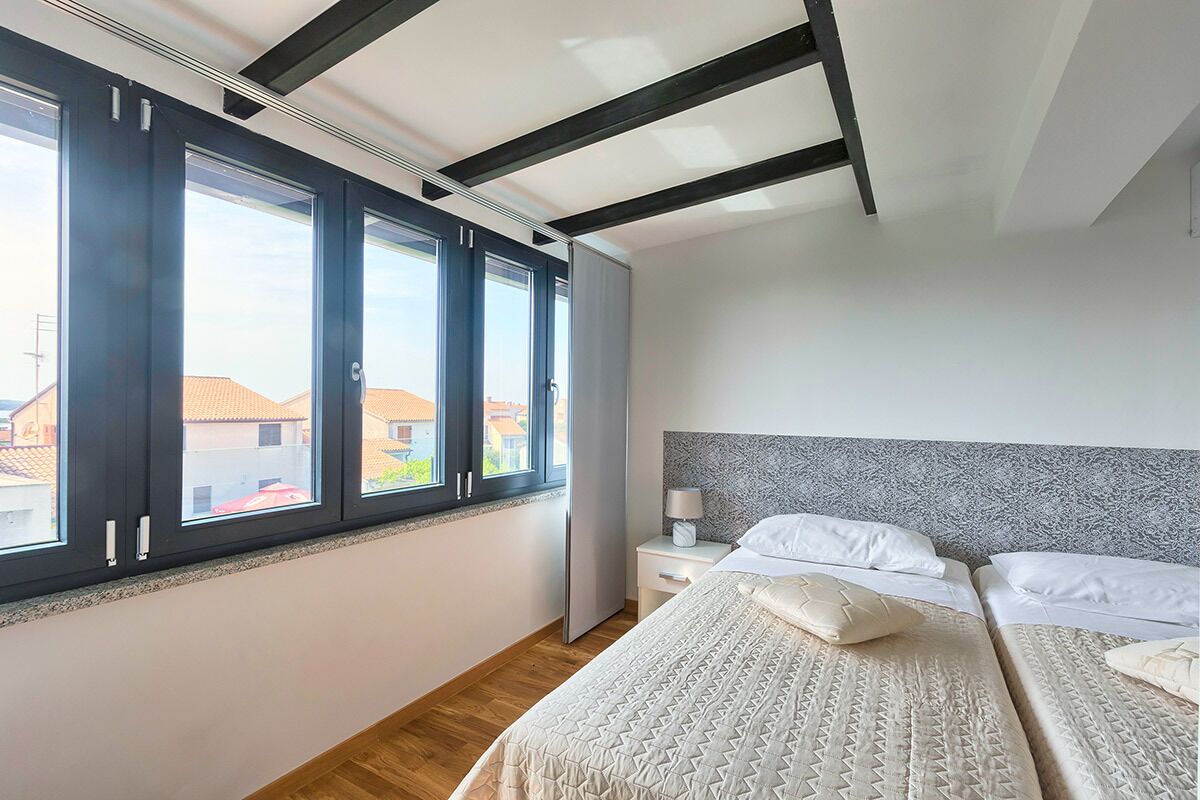 A bedroom with a large window, a bed with a patterned bedspread, a nightstand with a lamp, and a headboard with a textured design.