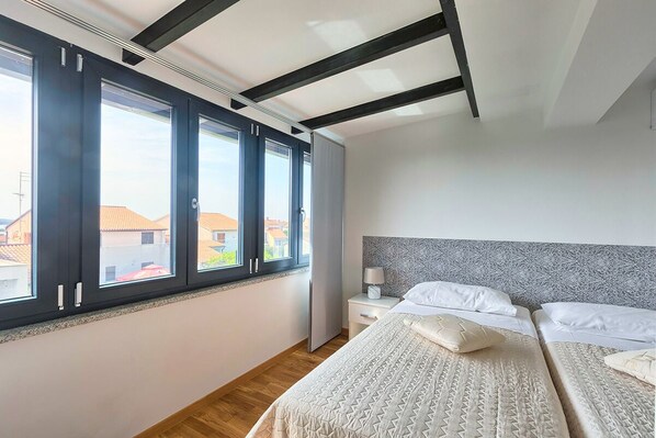 A bedroom with a large window, a bed with a patterned bedspread, a nightstand with a lamp, and a headboard with a textured design.