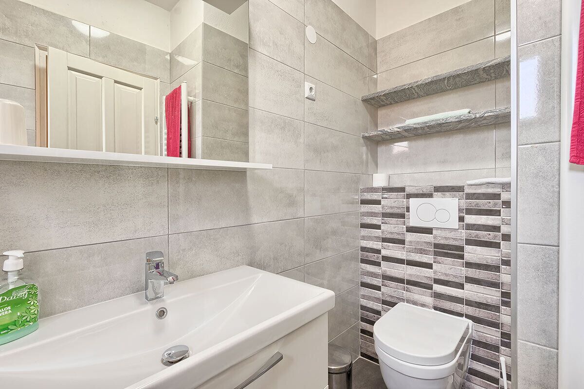 A bathroom with a white sink, a mirror, a toilet, and a shower area with tiled walls.