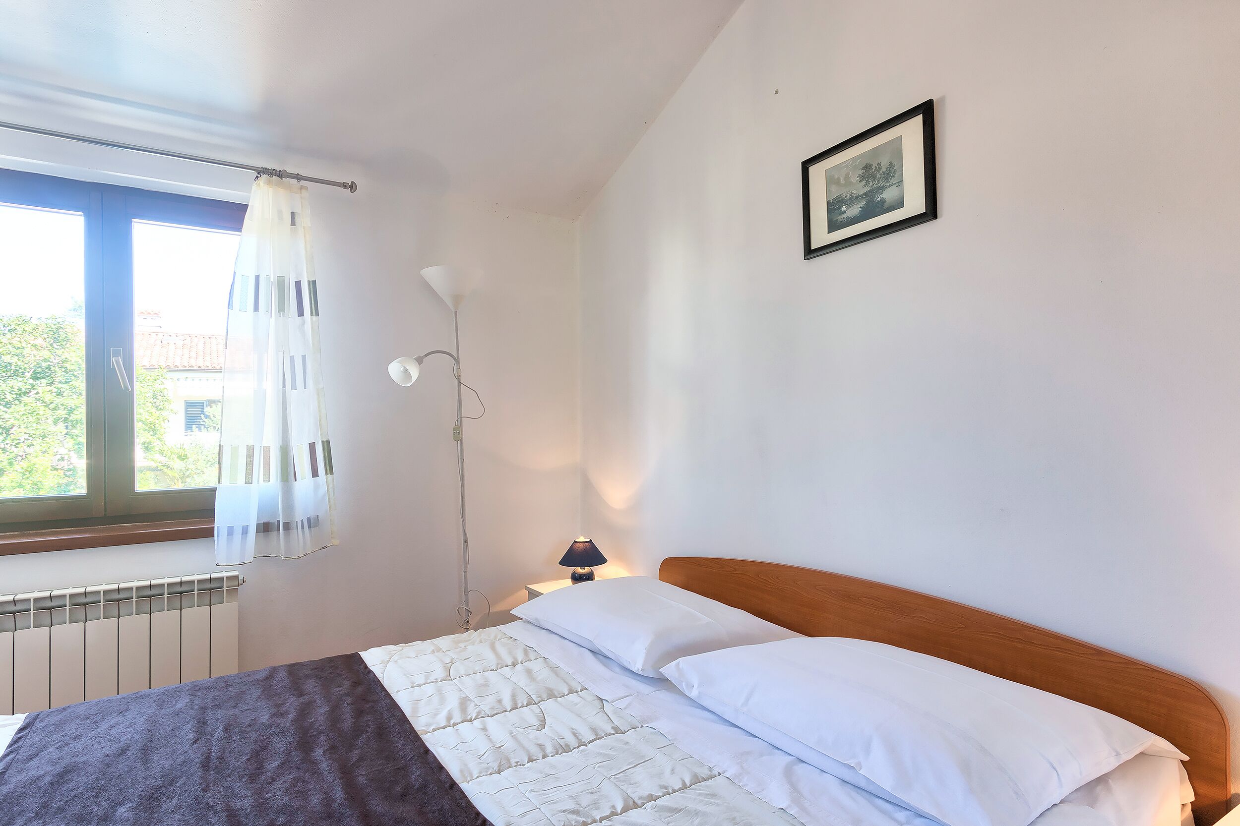 A bedroom with a bed, a window with curtains, a bedside lamp, and a framed picture on the wall.
