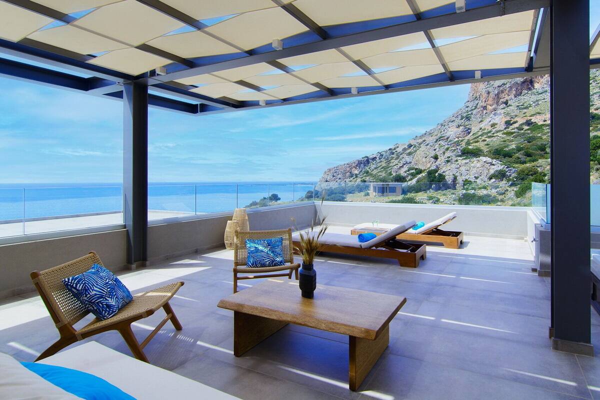 A spacious balcony with a view of the sea and a rocky hillside. There are wicker chairs, a wooden table, and a lounge chair with a cushion.