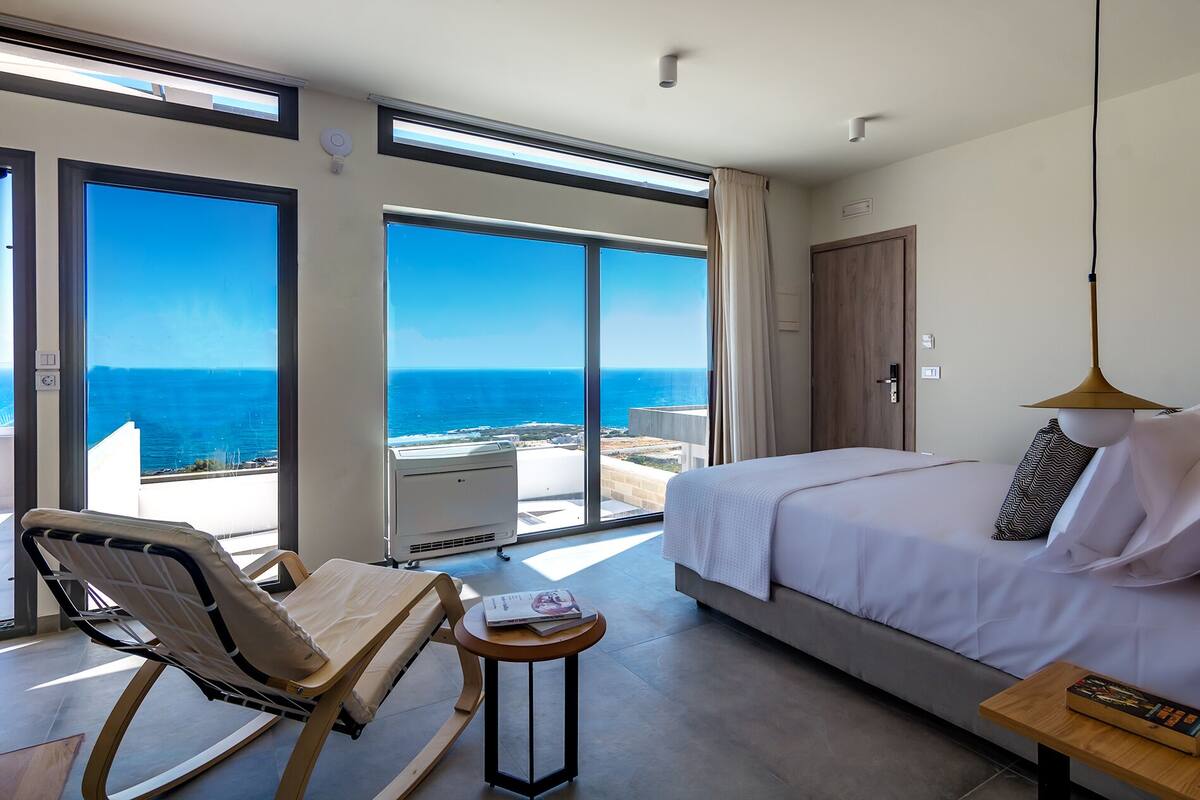 A hotel room with a bed, a chair, a small table, and a view of the ocean.