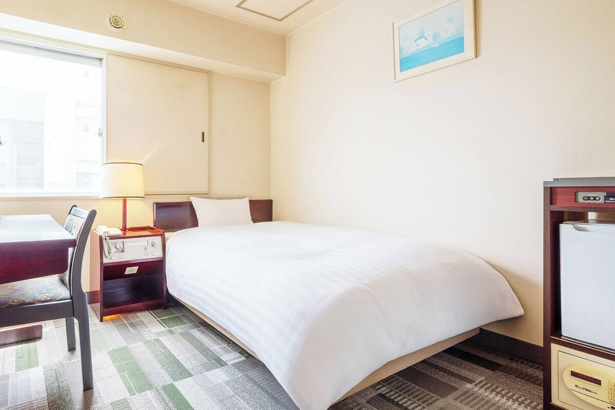 A hotel room with a bed, a desk with a lamp, a chair, a bedside table with a phone, and a picture on the wall.