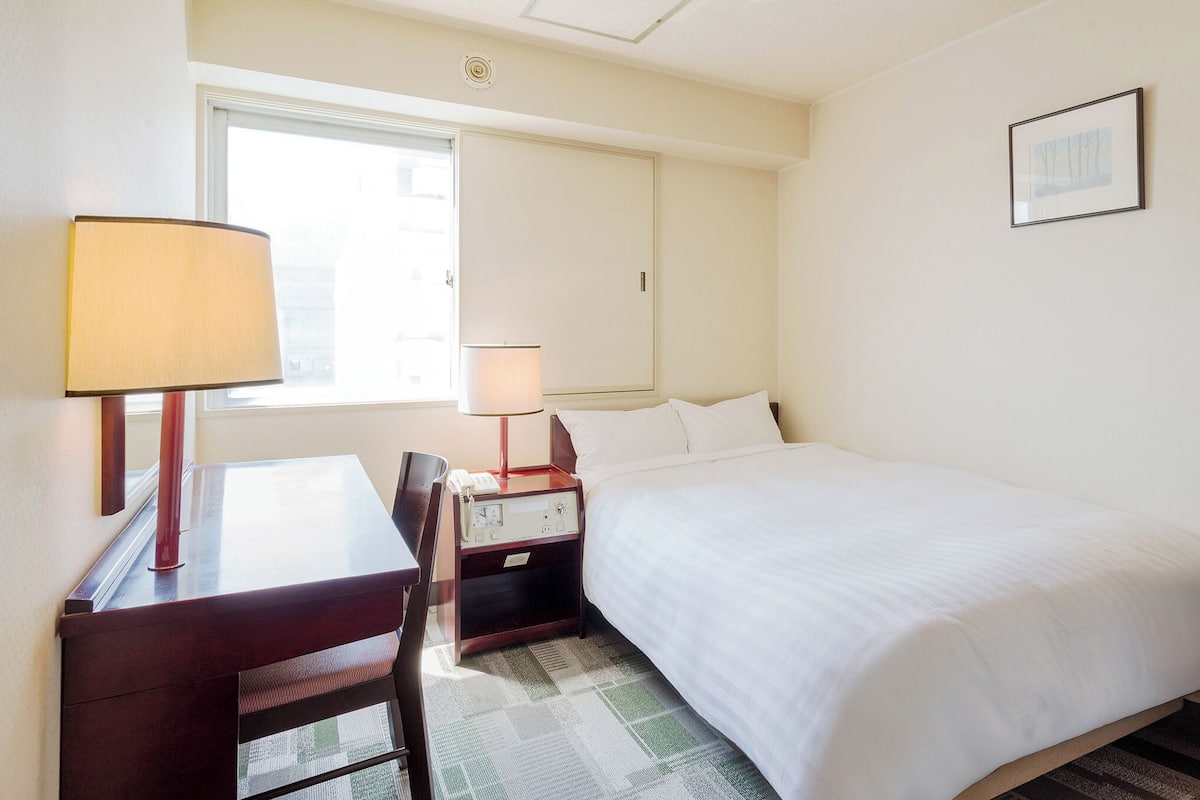 A hotel room with a bed, a desk with a lamp, a chair, a bedside table with a phone, and a picture on the wall.