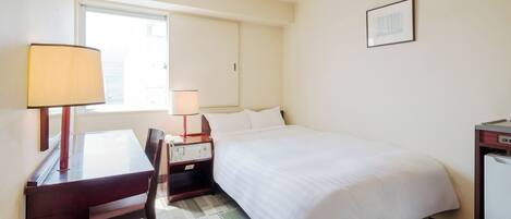 A hotel room with a bed, a desk with a lamp, a chair, a bedside table with a phone, and a picture on the wall.