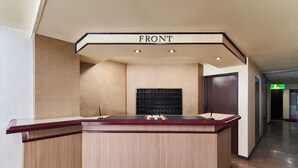 A reception desk with a sign reading "FRONT" above it, a black wall-mounted screen, and a wooden counter. - Hotel Paco Obihiro Chuo (Obihiro)