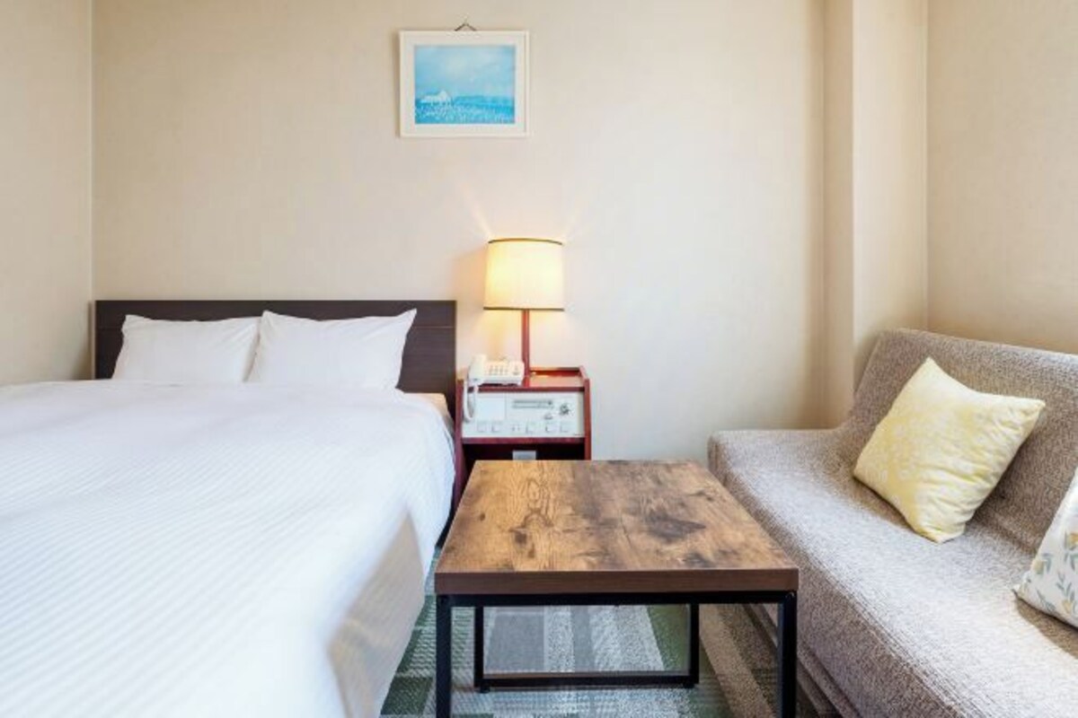 A hotel room with a bed, a sofa, a wooden coffee table, a nightstand with a lamp, and a painting on the wall.