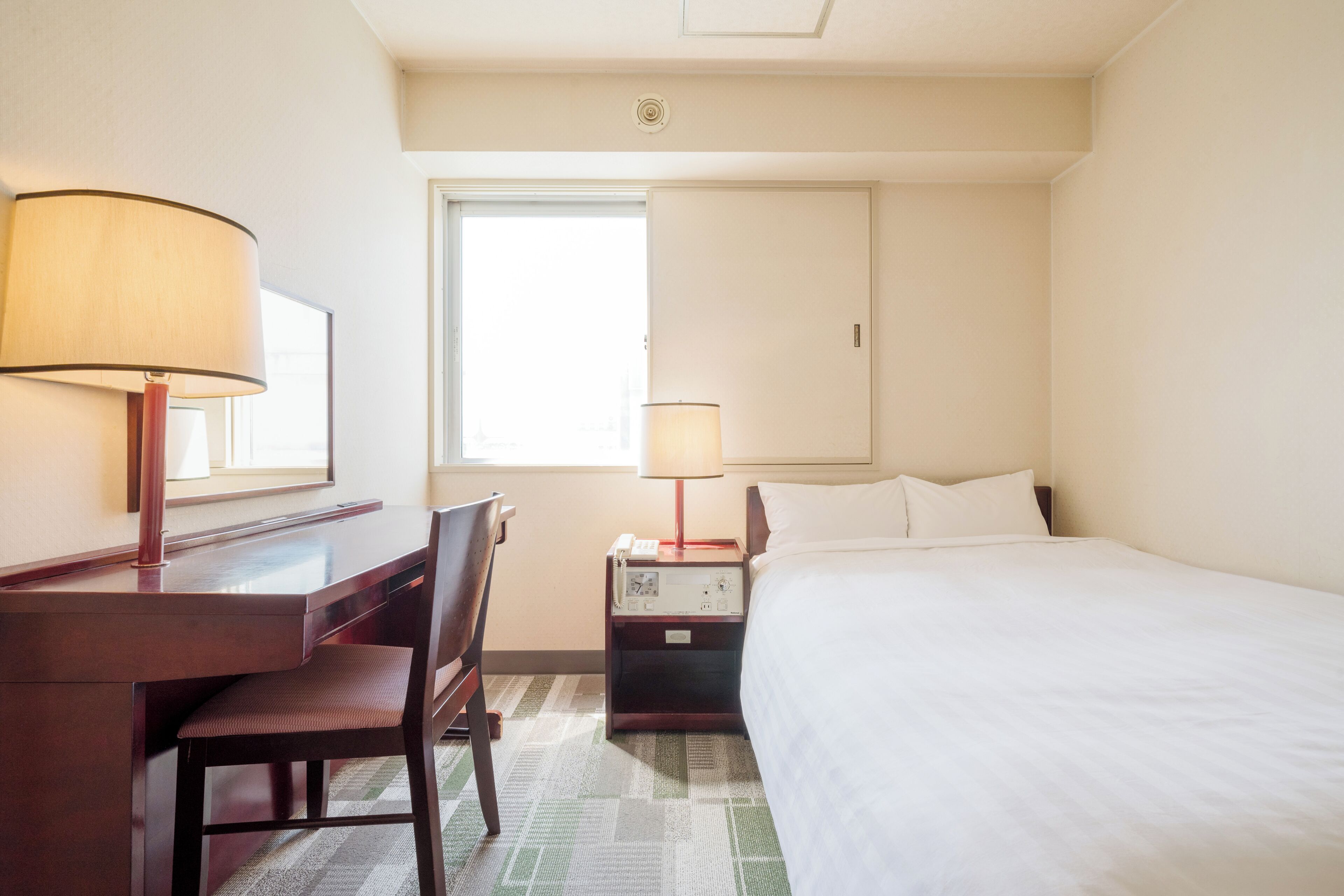 A hotel room with a bed, a desk, a chair, a lamp, and a nightstand with a phone.