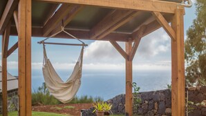 Uma pérgola de madeira com uma rede pendurada, com uma parede de pedra e uma área gramada ao fundo.