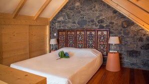 Quarto com teto de madeira, parede de pedra e cama com colcha branca e almofada decorativa.