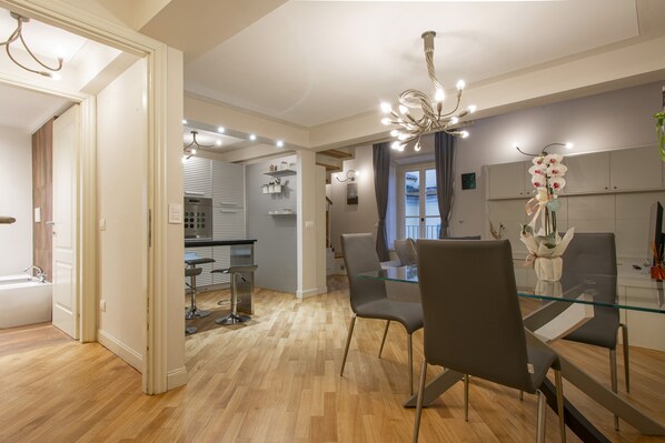 A modern kitchen with a built-in oven, a dining area with a glass table and chairs, and a chandelier.