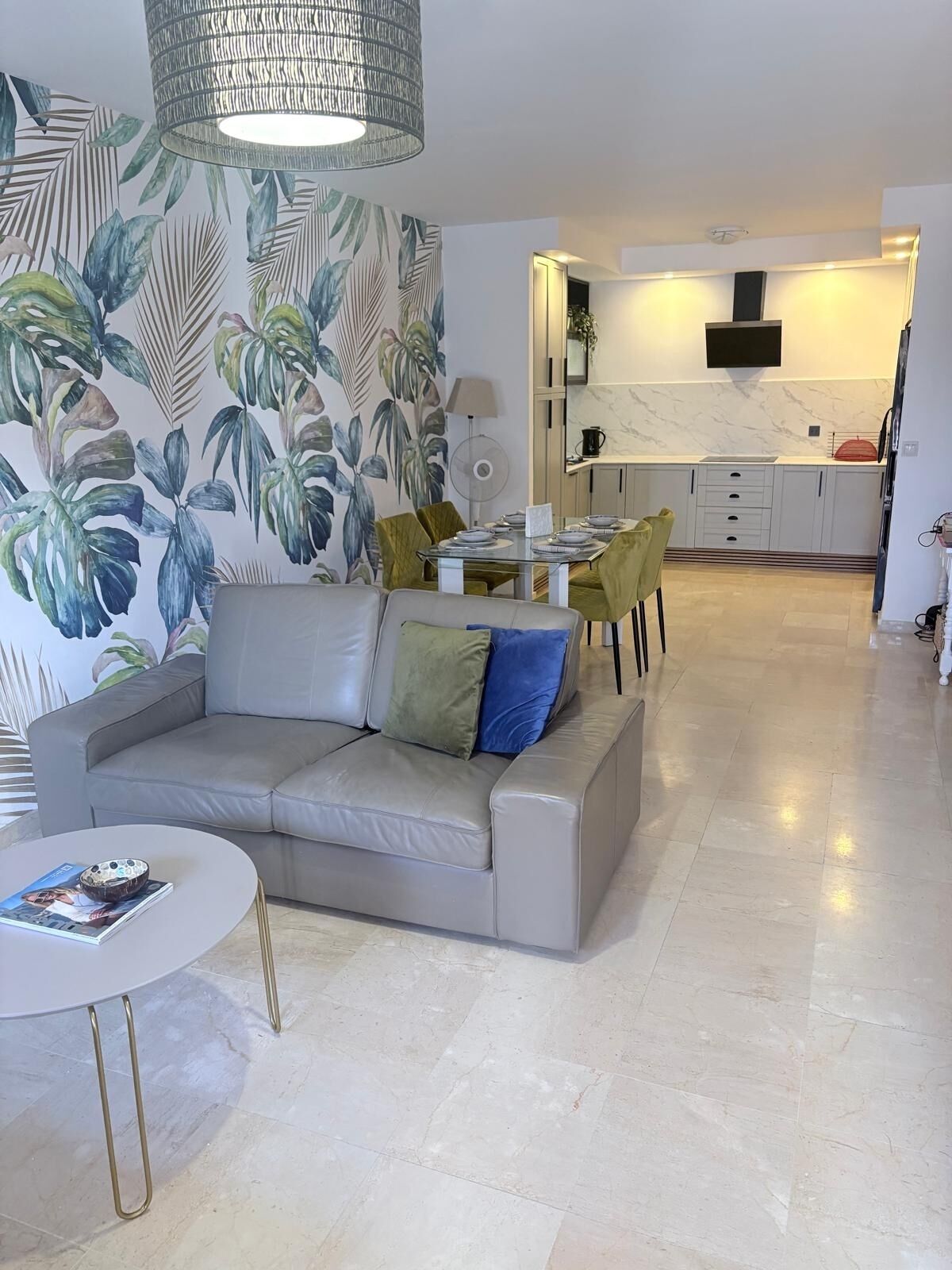 Een moderne woonkamer met een bank, een eettafel en stoelen. Er is een keuken met een marmeren aanrechtblad en een flatscreen televisie erboven.
