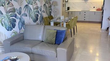 Een moderne woonkamer met een bank, een eettafel en stoelen. Er is een keuken met een marmeren aanrechtblad en een flatscreen televisie erboven.