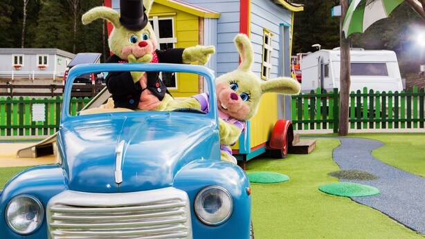 Une voiture bleue d’époque, avec deux personnages dans un décor ludique, une petite maison et une clôture verte.