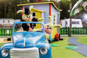 Une voiture bleue d’époque, avec deux personnages dans un décor ludique, une petite maison et une clôture verte.