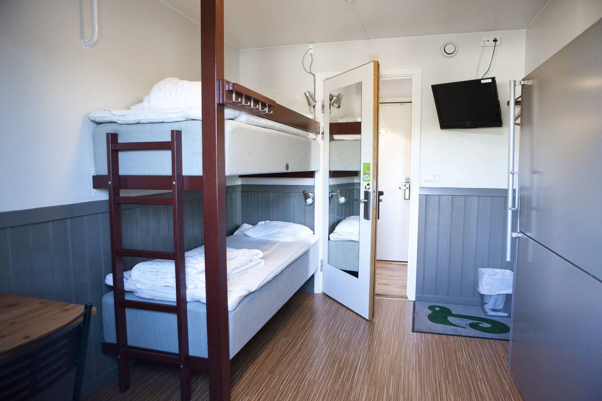 Een slaapkamer met stapelbedden, een houten trapje, een klein tafeltje en een deur naar een badkamer met een spiegel en toilet.