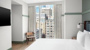Hotelkamer met een groot bed, een stoel, een aan de muur bevestigde televisie en uitzicht op stadsgebouwen.