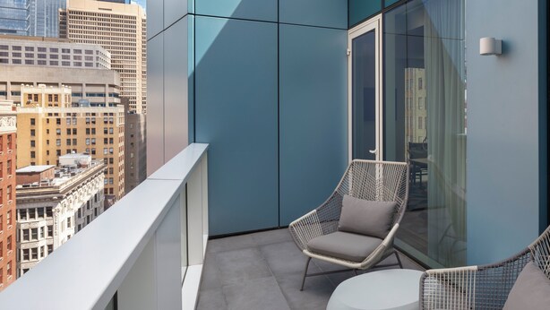 Een balkon met rieten stoelen en een kleine ronde tafel, met uitzicht op een stadsgezicht met moderne gebouwen.