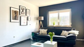 Un salon moderne avec un canapé en cuir noir, une table basse en verre et des œuvres d’art encadrées accrochées au mur.