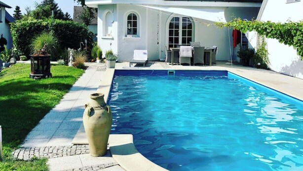 Une grande piscine bordée de pierres, une maison blanche dotée d’une tourelle et un jardin bien entretenu.
