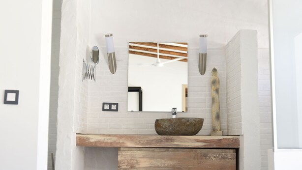 A bathroom with a white brick wall, a wooden vanity, a large mirror, and a stone basin.