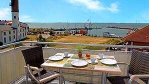 Une table de balcon avec des assiettes, des tasses et une plante, donnant sur un port où l’on peut voir des bateaux et un phare.