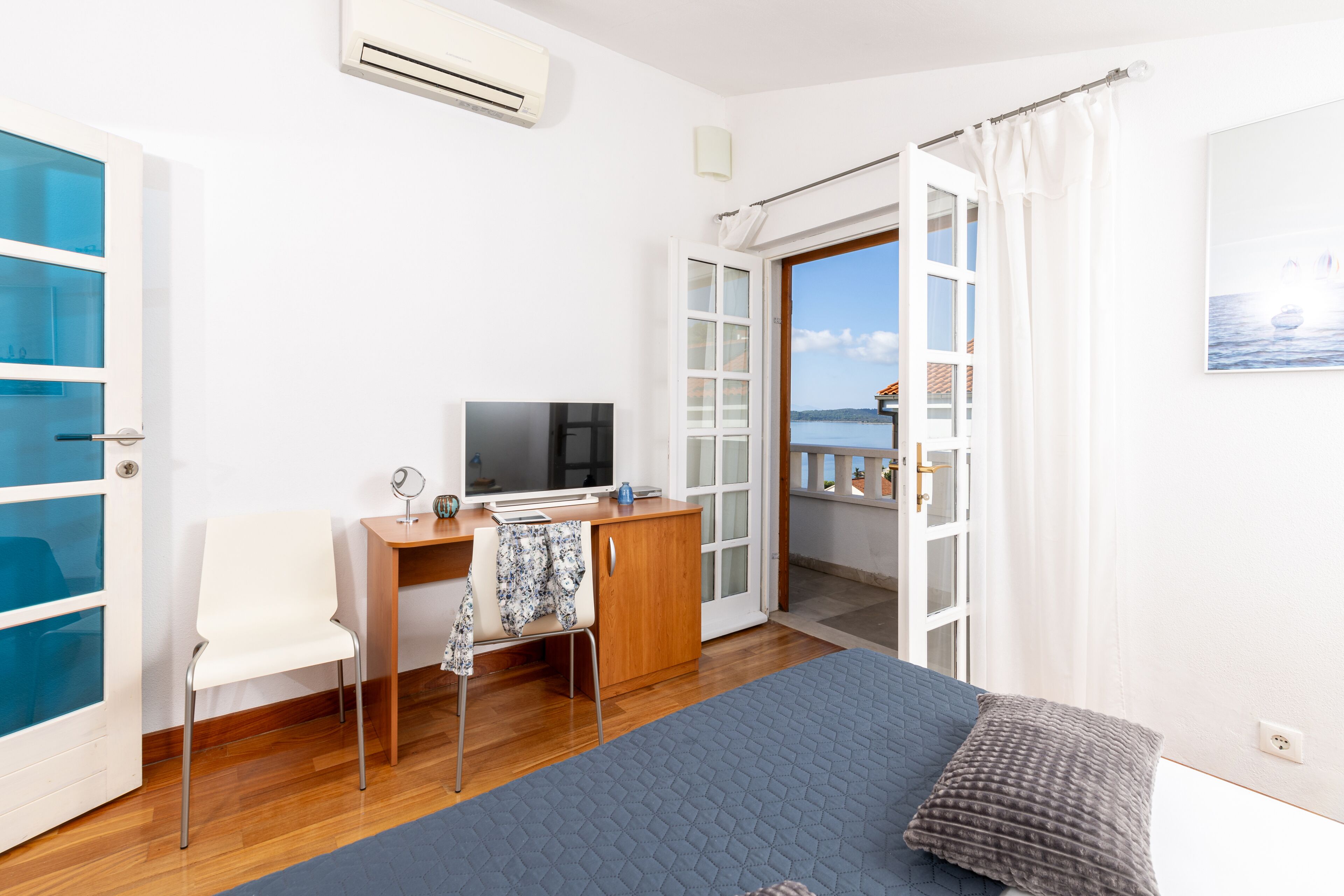 Un dormitorio con cama, escritorio, silla, televisor y vista al mar a través de una puerta abierta.