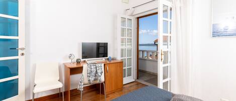 Un dormitorio con cama, escritorio, silla, televisor y vista al mar a través de una puerta abierta.
