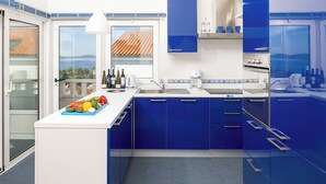 A modern kitchen with blue cabinets, a white countertop, and a view of the sea through a large window.