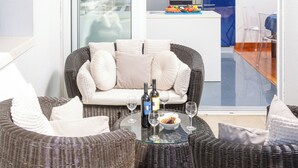 A modern living area with wicker furniture, a glass table, wine bottles, and a view of the kitchen.