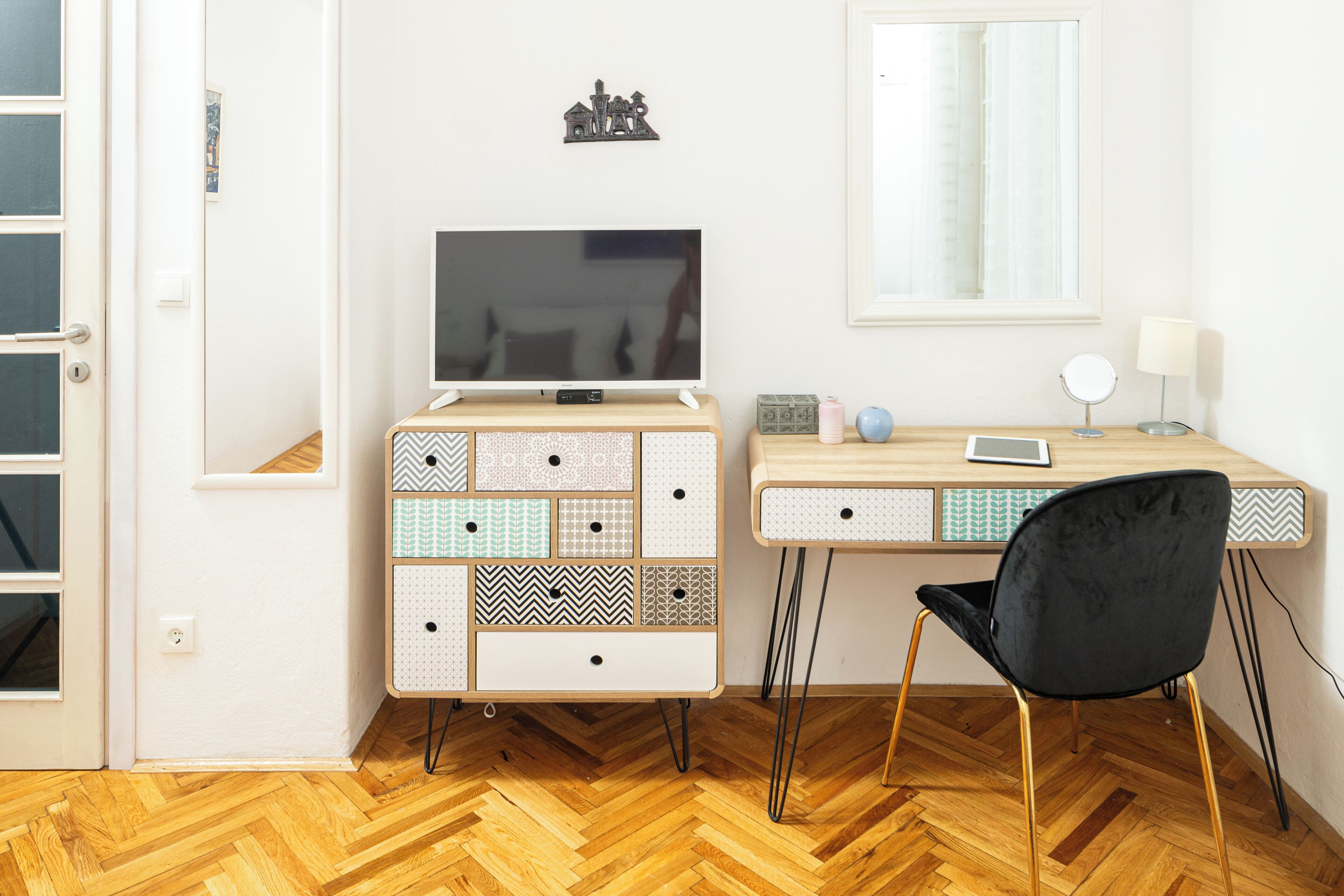 A room with a wooden herringbone floor, a white wall, a TV on a wooden cabinet, a desk with a chair, and a window with a curtain.
