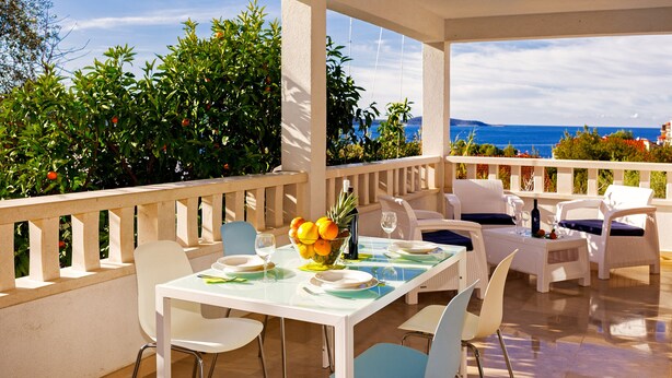 A terrace with white furniture, a table set with plates and glasses, and a bowl of fruit.