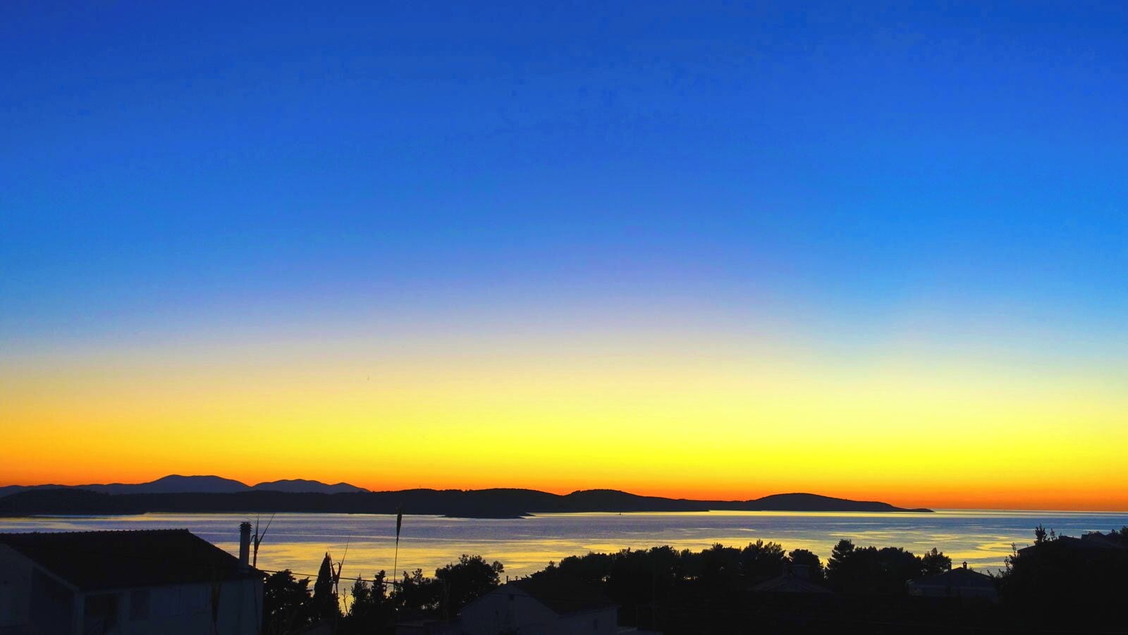 Eine Küstenlandschaft mit Sonnenuntergang über dem Wasser, Häusersilhouetten und in der Ferne Hügel.