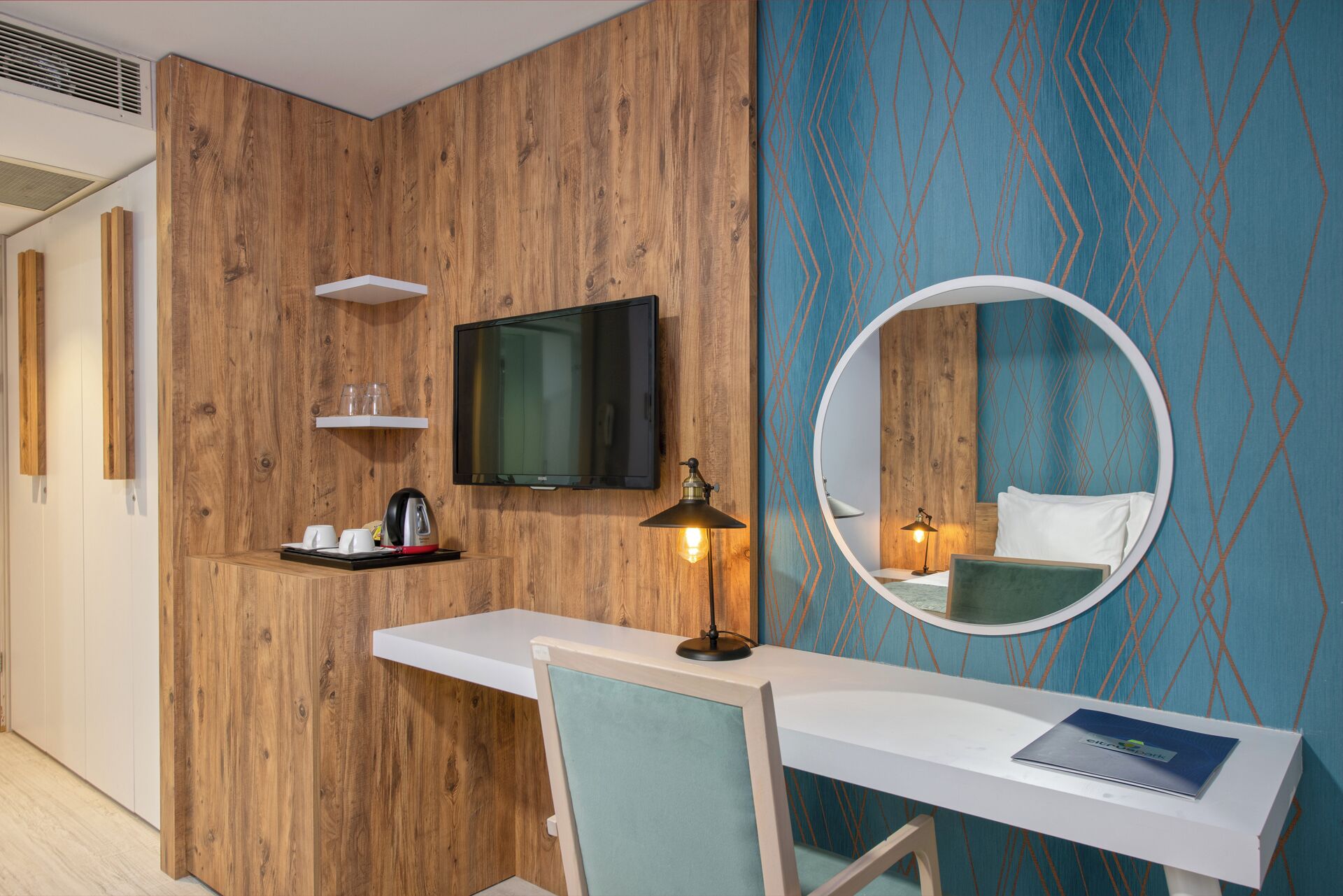 A hotel room with a wooden accent wall, a flat-screen TV, a round mirror, a desk with a chair, and a bookshelf.