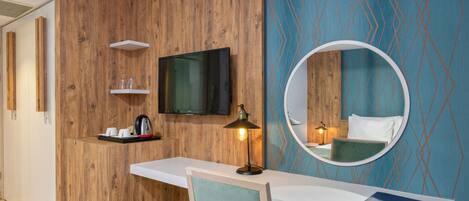 A hotel room with a wooden accent wall, a flat-screen TV, a round mirror, a desk with a chair, and a bookshelf.