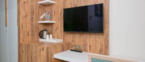 A modern room with a wooden accent wall, a mounted TV, a white shelf with a water bottle, a kettle, and a remote control.