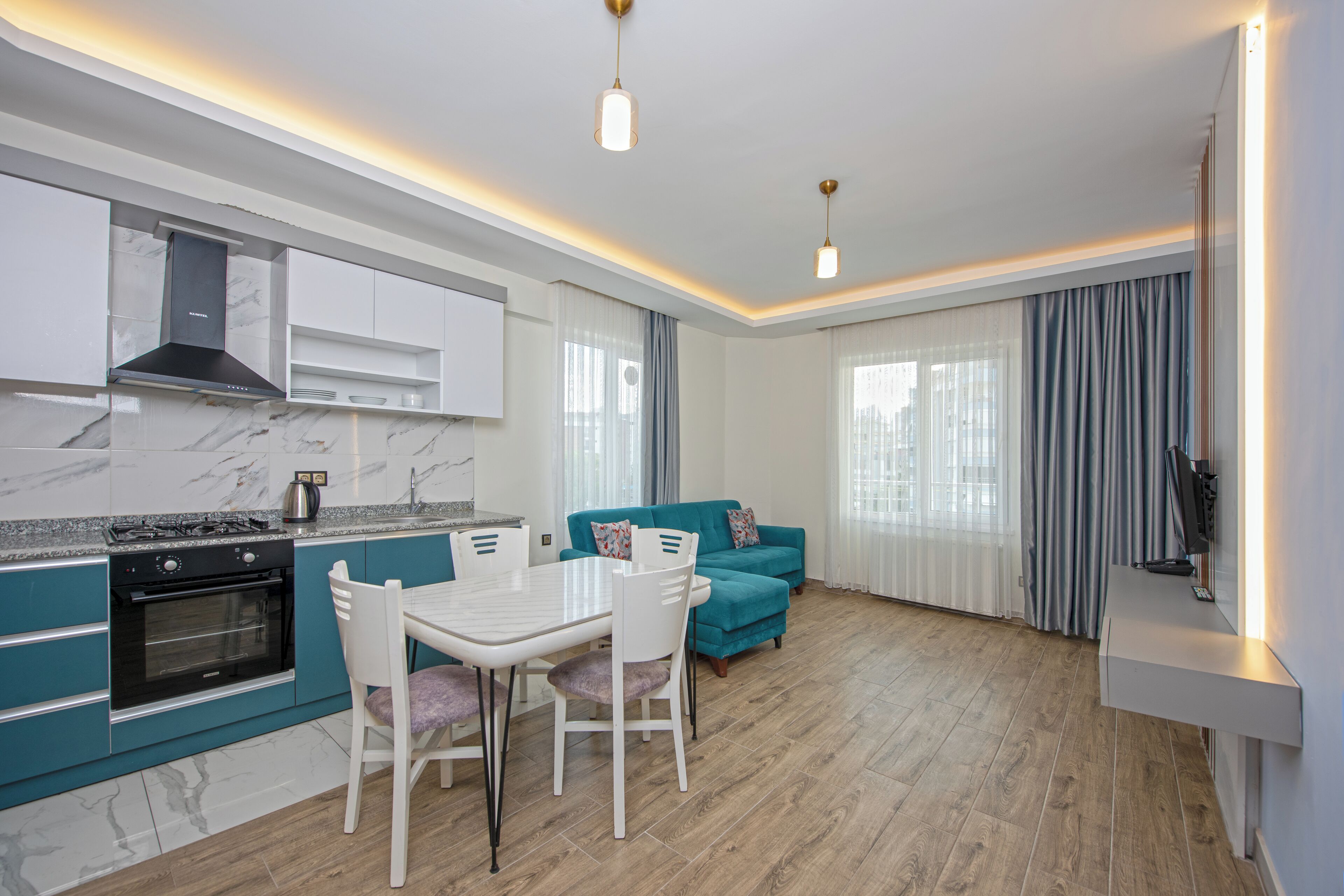 Una cocina moderna con estufa y horno integrados, una mesa de comedor con sillas, un sofá color turquesa y un televisor de pantalla plana montado en la pared.