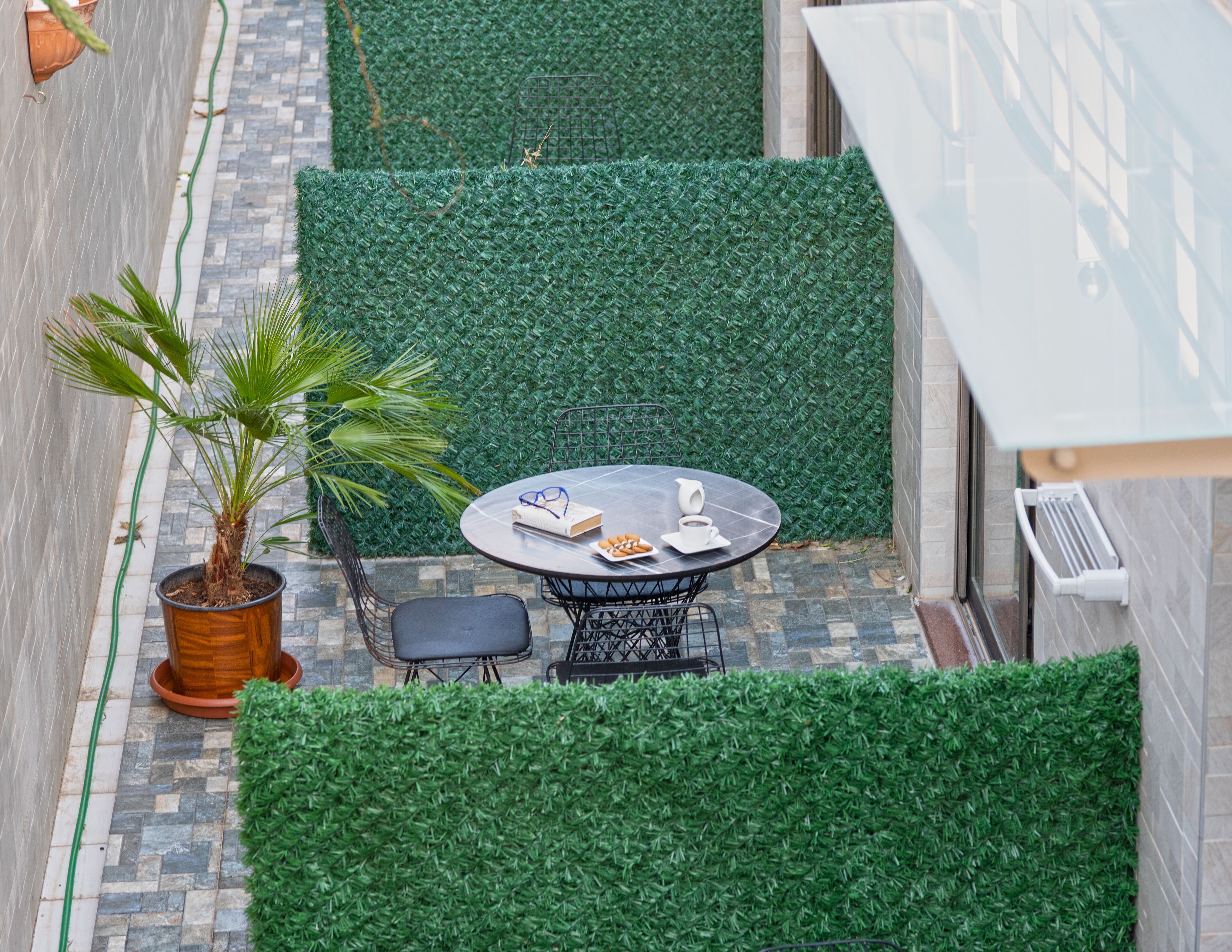 A small outdoor seating area with a round table and chairs, surrounded by green artificial hedges and a potted palm plant.