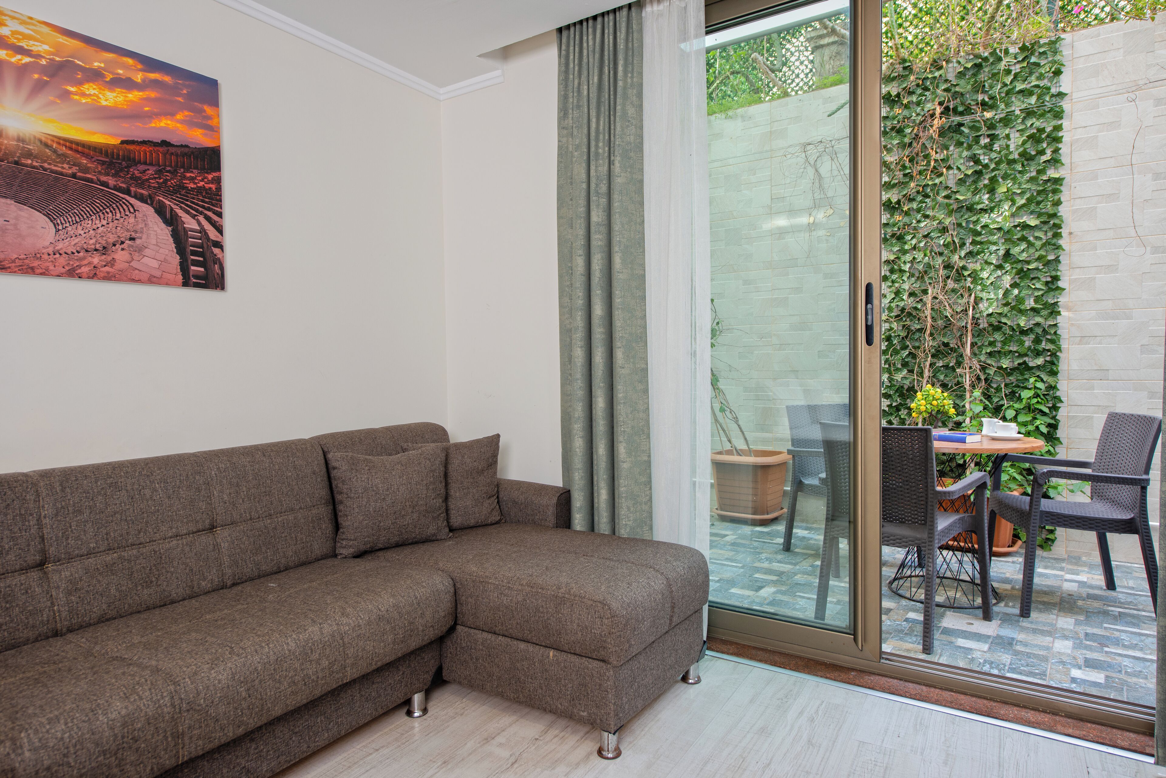 A living room with a sectional sofa, a glass door leading to a patio with a table and chairs, and a wall-mounted painting.